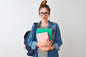 A student with glasses is standing and looking at the camera with their notebooks and backpack.