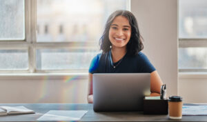 A student sitting at a table with her laptop and smiling at the camera.