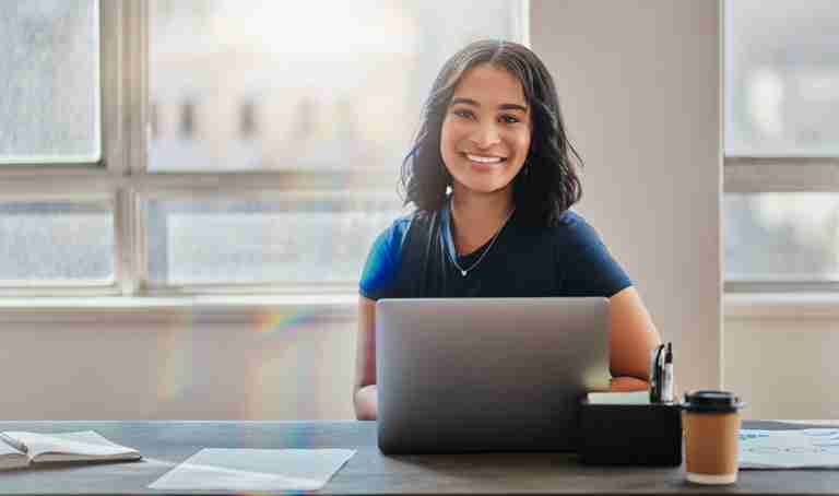 A student sitting at a table with her laptop and smiling at the camera.