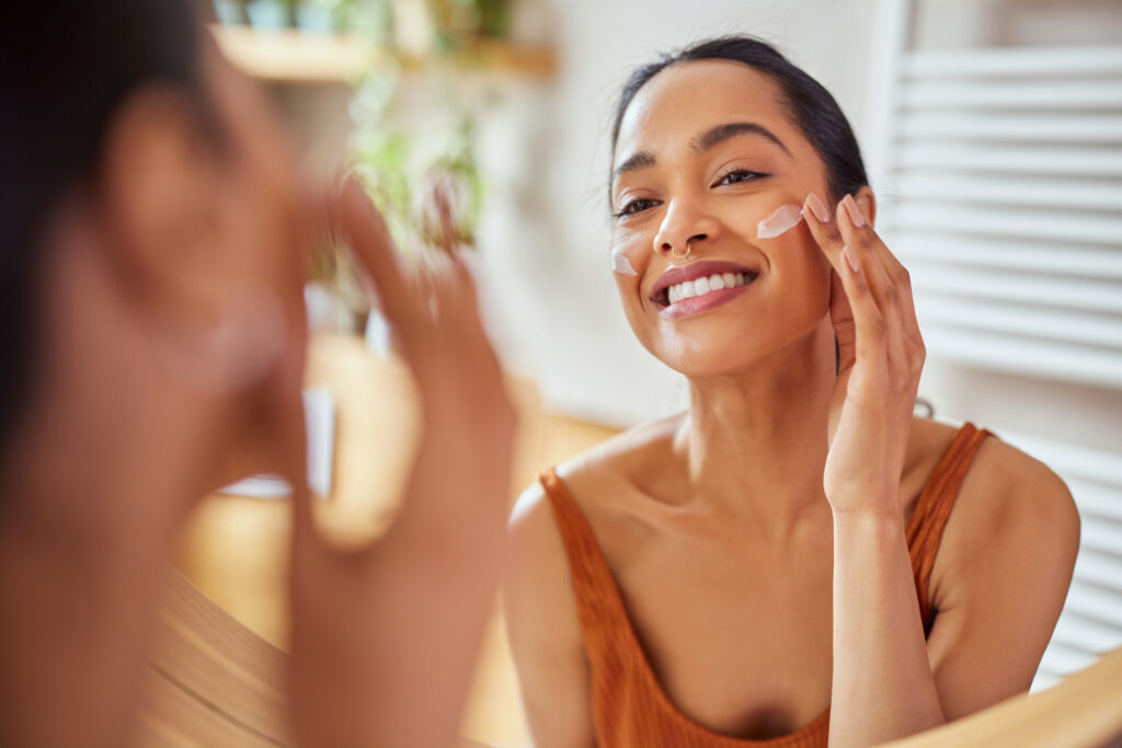 A women smiling in the mirror at herself and applying face cream to her cheeks.
