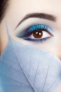 A closeup of a woman's eye with blue and green eyeshadow and eyeliner. She has a bluish leaf held up to her cheek.