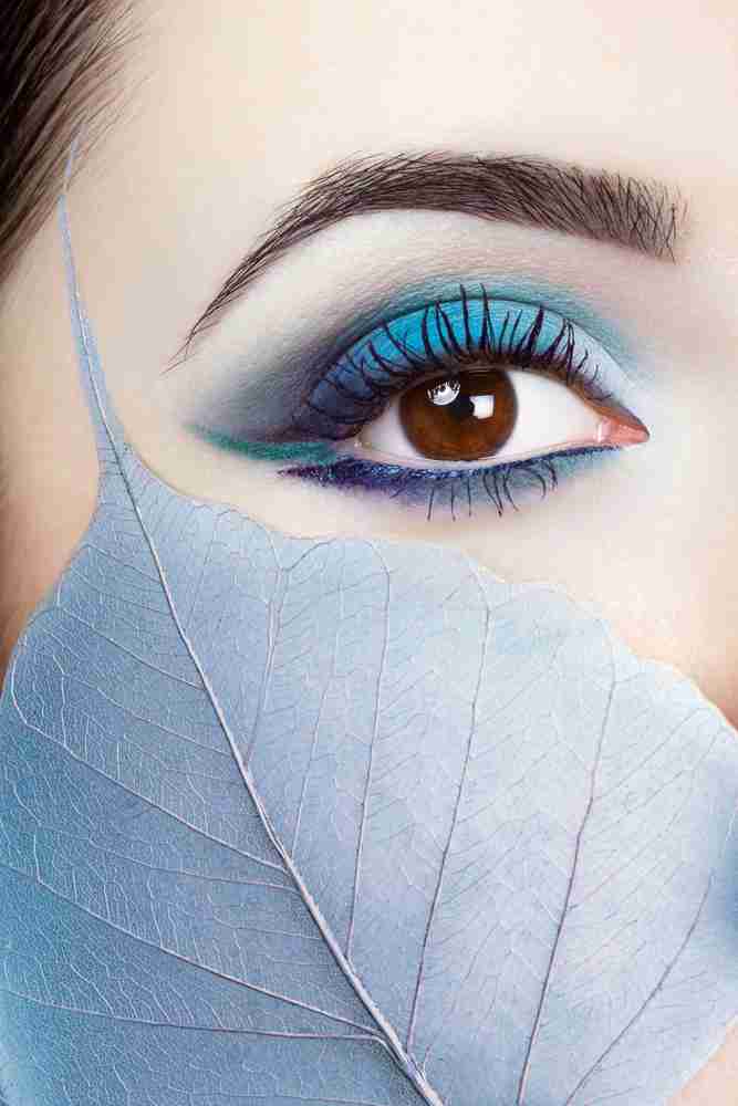 A closeup of a woman's eye with blue and green eyeshadow and eyeliner. She has a bluish leaf held up to her cheek.