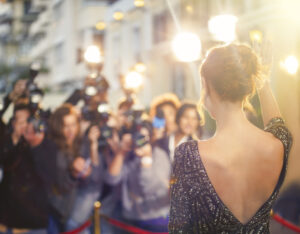A woman on a red carpet in an event dress with her back to the camera. She is smiling and facing a crowd of papparazi.