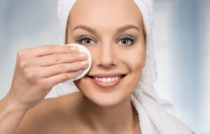 A profile view of a woman smiling at the camera and removing her makeup with a makeup pad. She has a towel on her head as well.