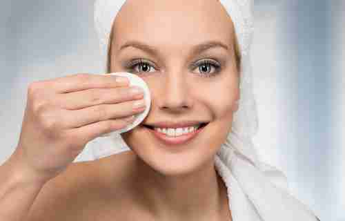 A profile view of a woman smiling at the camera and removing her makeup with a makeup pad. She has a towel on her head as well.