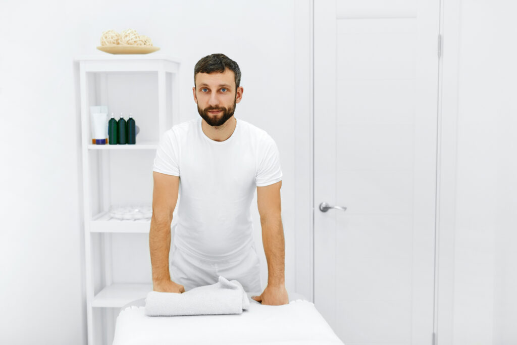 A massage therapist in a white room dressed in white smiling at the camera and leaning on a massage table.