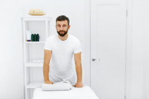 A massage therapist in a white room dressed in white smiling at the camera and leaning on a massage table.