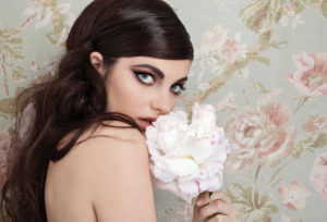 Model with long brunette hair with smokey makeup featuring heavy winged eyeliner, holding a flower in front of a floral-themed wall. 