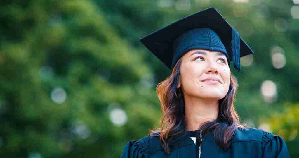 Beauty school graduate smiling in cap and gown outdoors.